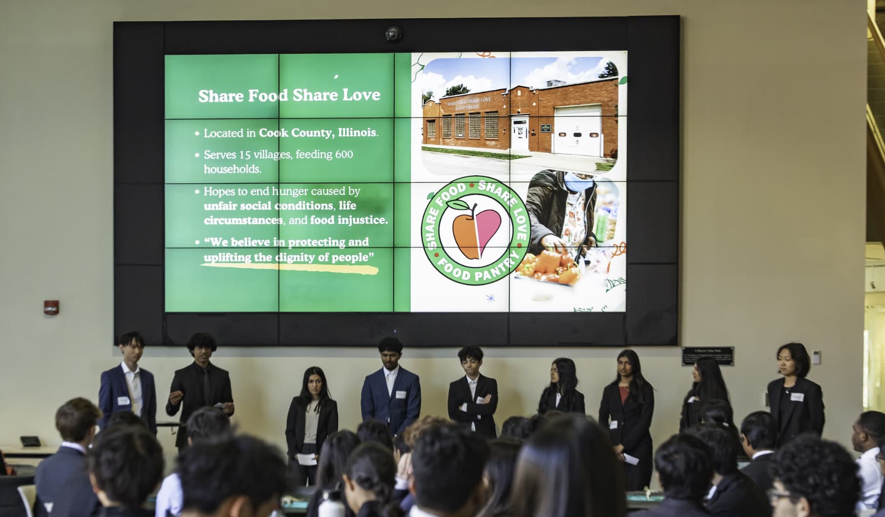 A Hack the Future team presenting to the club in front of a video wall showing its nonprofit, the Share Food Share Love food pantry