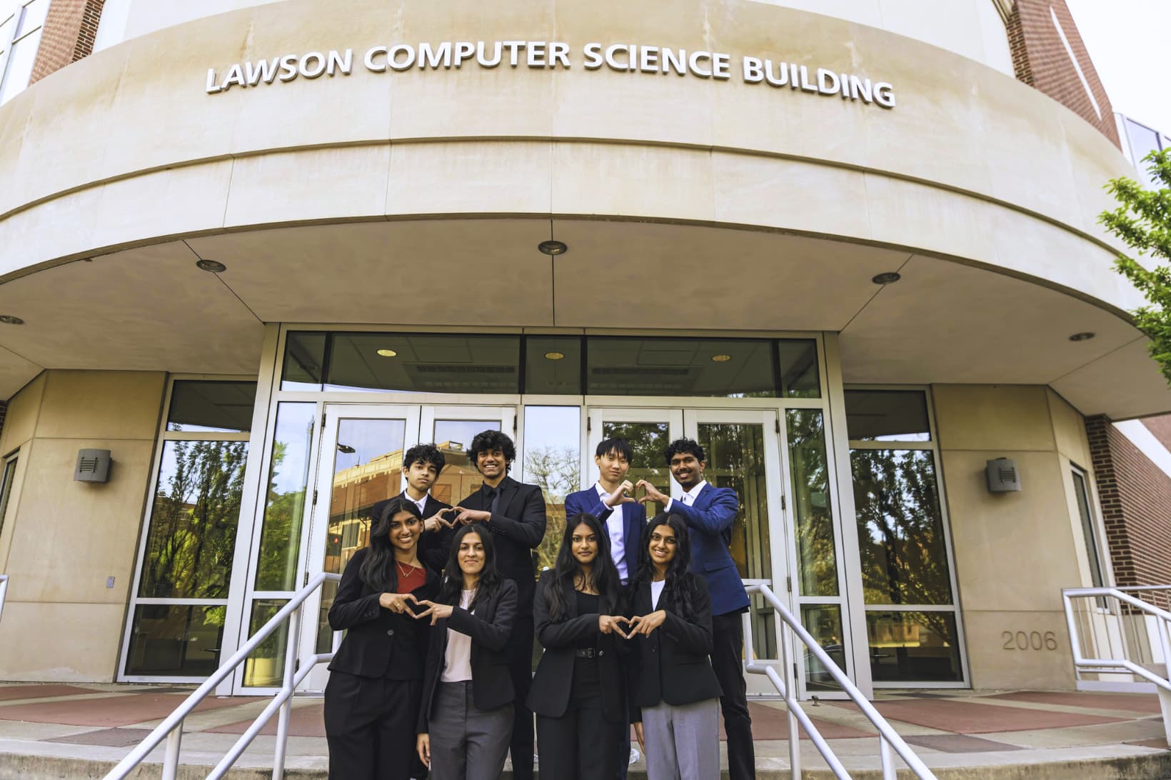 Eight Hack the Future members in business attire making heart shapes with their hands on the steps of the Lawson Computer Science Building